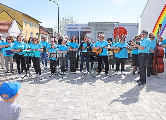 Eine Gruppe von Musikern in blauen T-Shirts spielt Gitarren auf einer gepflasterten Straße mit Regenbogenhintergrund und einer zuschauenden Menge.