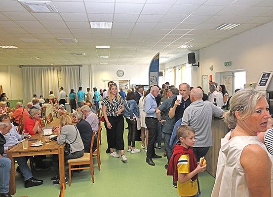 Ein überfüllter Speisebereich mit Tischen und Stühlen. Die Menschen essen, unterhalten sich und stehen herum. Einige tragen Freizeitkleidung, Brillen und Turnschuhe. Eine Uhr ist an der Wand montiert.