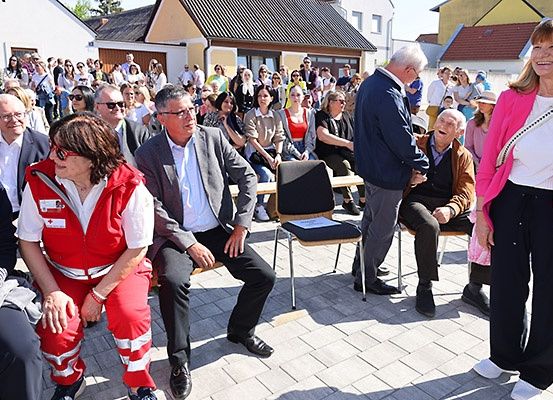 Eine Menschenmenge, darunter ein Mann in einem grauen Anzug und eine Frau in einer roten Jacke, versammelt sich auf einem Außenbereich, wahrscheinlich für eine Veranstaltung.