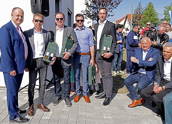 Mehrere Männer in Anzügen und Krawatten halten Weinflaschen in einer Gruppenaufnahme. Der Mann in der Mitte trägt eine Sonnenbrille. Sie posieren für ein Foto draußen.