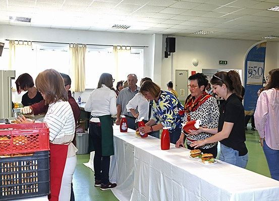 Eine Gruppe von Menschen steht in einer Schlange an einem langen Tisch, möglicherweise in einer Cafeteria. Der Tisch ist mit einem weißen Tuch bedeckt, und es befinden sich mehrere Gegenstände darauf. Einige Menschen tragen eine Brille, und eine Person trägt eine Armbanduhr. Auf der linken Seite steht ein roter Korb. Hinter dem Tisch befindet sich eine Glastür mit einer Gardine.