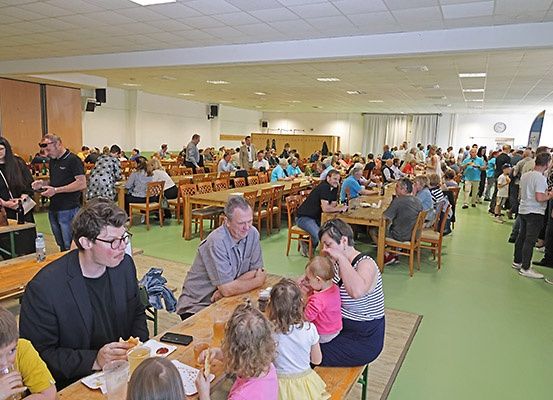 Ein überfüllter Raum mit mehreren Personen, die an Tischen sitzen, andere stehen und eine Frau ein Baby hält, während ein Mann in der Nähe einer Tür isst.