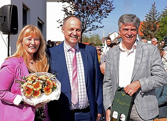 Drei Personen stehen lächelnd eng beieinander im Freien. Die Frau hält einen Blumenstrauß, und die Männer halten eine Geschenkbox und nichts. Dahinter stehen und gehen Leute. Bäume und Gebäude sind im Hintergrund.