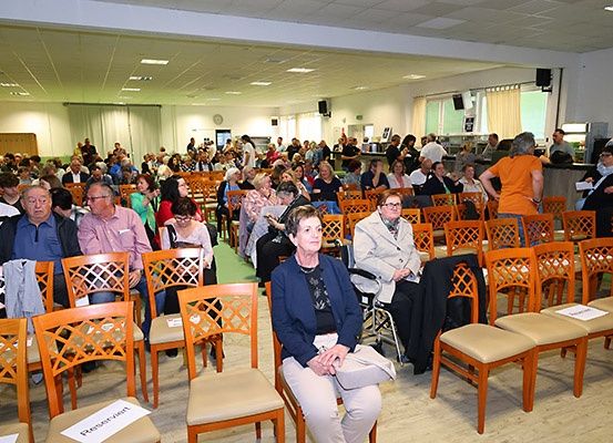 Ein Saal voller Menschen, die in Reihen aus Holzstühlen sitzen, einige mit Brille, einem grünen Teppich auf dem Boden und einer Uhr an der Wand.