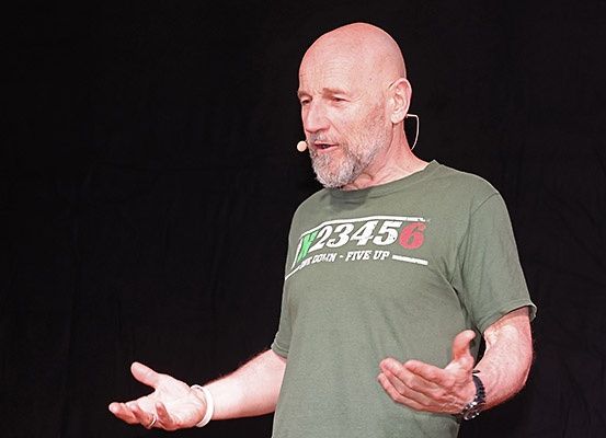 Ein kahlköpfiger Mann in einem grünen T-Shirt mit weißem Text spricht in ein Mikrofon auf der Bühne und gestikuliert mit den Händen.