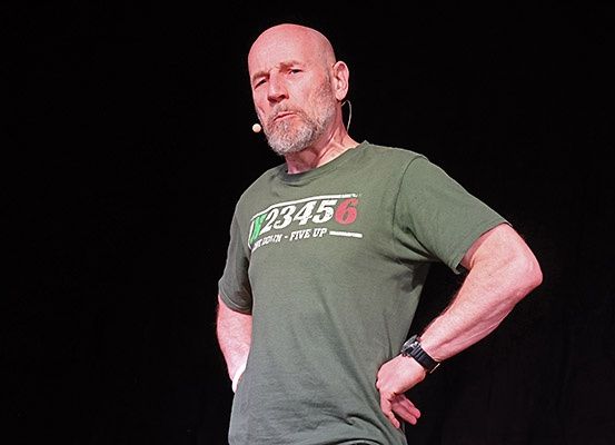 Ein kahlköpfiger Mann mit einem Mikrofon am Ohr, der ein grünes T-Shirt trägt, steht mit den Händen in den Hüften auf der Bühne.