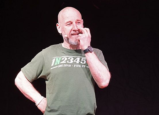 Ein kahlköpfiger Mann mit einer grünen T-Shirt, auf dem 'IN2345' steht, und einer Uhr steht auf der Bühne und berührt sein Gesicht.
