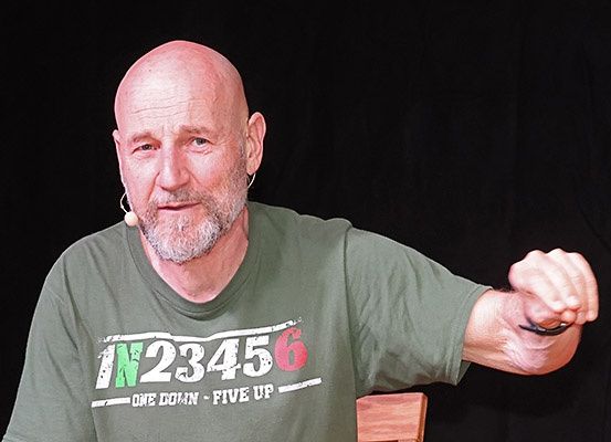 Ein älterer kahlköpfiger Mann in einem grünen T-Shirt mit den Worten 'N23456' und 'EINER RUNTER FÜNF OBEN' spricht, während er sich auf einen Holztisch lehnt.