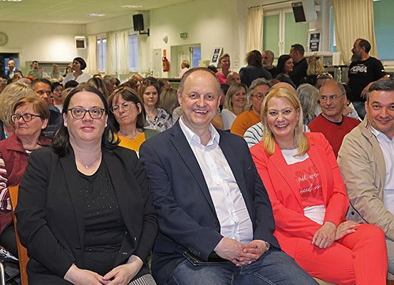 Eine Gruppe von Menschen sitzt in Stühlen in einem Raum mit einem lächelnden Mann in der Mitte, einer Frau in einer roten Jacke und einem Mann in einem weißen Hemd.
