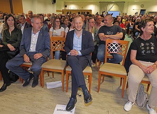 Eine Gruppe von Menschen sitzt auf Stühlen, wahrscheinlich bei einer Versammlung. Ein Mann in der Mitte lächelt und scheint für ein Foto zu posieren.
