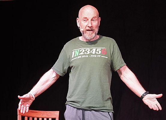 Ein älterer Mann trägt ein grünes T-Shirt mit IN23456 und ONE DOLLAR FIVE UP aufgedruckt, er steht auf einer Bühne.
