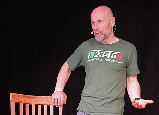 Ein kahlköpfiger Mann steht auf einer Bühne, trägt ein grünes T-Shirt mit einem Design und ein Mikrofon und spricht zu einem Publikum.