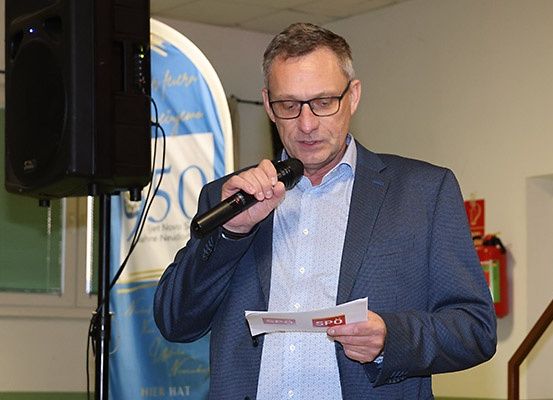 Ein Mann mit Brille und Anzug spricht in ein Mikrofon und hält ein Papier in der anderen Hand. Hinter ihm steht ein Lautsprecher auf einem Ständer, und neben ihm ist ein Banner angebracht. An der Wand rechts ist ein Feuerlöscher angebracht.
