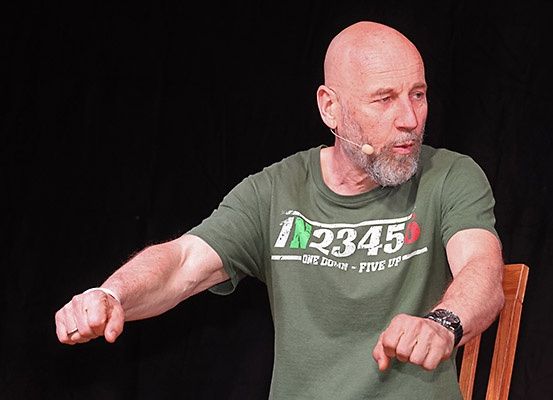 Ein kahlköpfiger Mann in einem grünen T-Shirt mit Text und Zahlen sitzt auf einem Holzstuhl und gestikuliert mit den Händen, während er ein Mikrofon-Headset trägt.