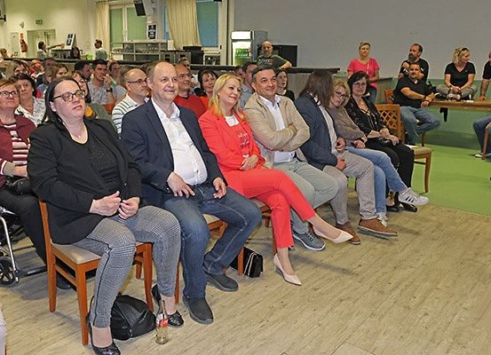 Eine Gruppe von Menschen sitzt in einem Raum, einige mit Brille, andere in formeller Kleidung. Ein Mann im Anzug lächelt, während er mit einer Frau in einem roten Outfit sitzt. Hinter ihnen steht eine Person in einem rosa Shirt.
