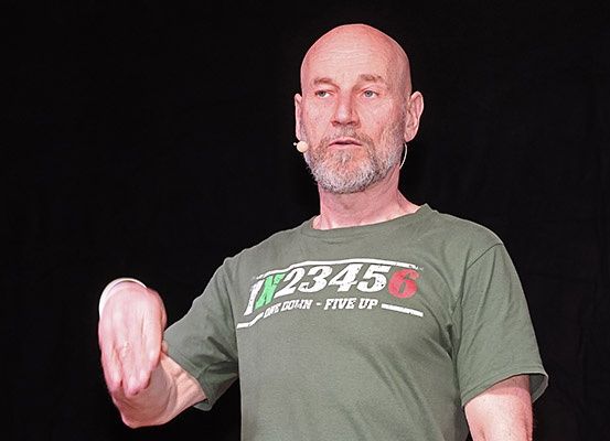 Ein kahlköpfiger Mann in einem grünen T-Shirt mit weißer und roter Schrift, möglicherweise bei einer Rede. Er trägt ein Mikrofon am Ohr.