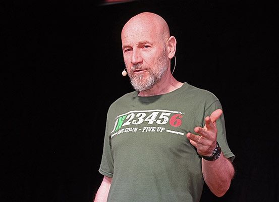 Ein kahlköpfiger Mann steht auf der Bühne und trägt ein grünes T-Shirt mit weißem Text. Er gestikuliert während des Sprechens und trägt ein verkabeltes Mikrofon.