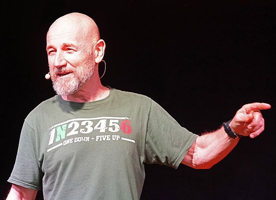 Ein kahlköpfiger Mann in einem grauen T-Shirt spricht, zeigt mit der rechten Hand und trägt ein Mikrofon-Ohrstück.