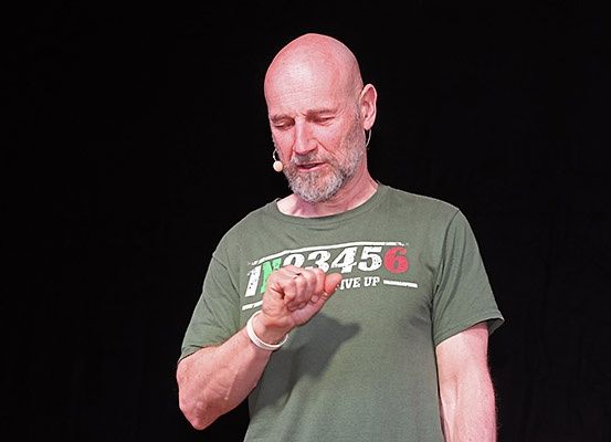 Ein kahlköpfiger Mann in einem grünen T-Shirt hält eine Rede auf der Bühne und hält ein kleines Objekt in der Hand. Er trägt eine Armband und einen verkabelten Ohrhörer.