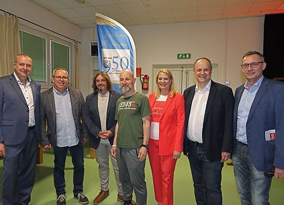 Sechs Erwachsene, gekleidet in formeller und halbformeller Kleidung, stehen lächelnd zusammen für ein Foto. Ein blauer Banner mit '950' und einem Logo ist hinter ihnen. Ein Feuerlöscher ist an der Wand montiert.