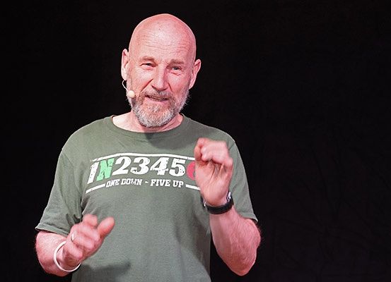 Ein kahlköpfiger Mann in einem grünen T-Shirt mit Text und einem Mikrofon am Ohr lächelt und gestikuliert, während er auf der Bühne spricht.