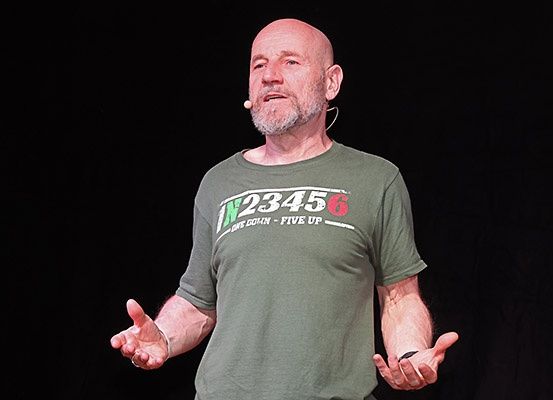 Ein kahlköpfiger Mann mit Bart, der ein grünes T-Shirt mit weißem Text trägt, steht auf der Bühne und spricht, während er mit den Händen gestikuliert.
