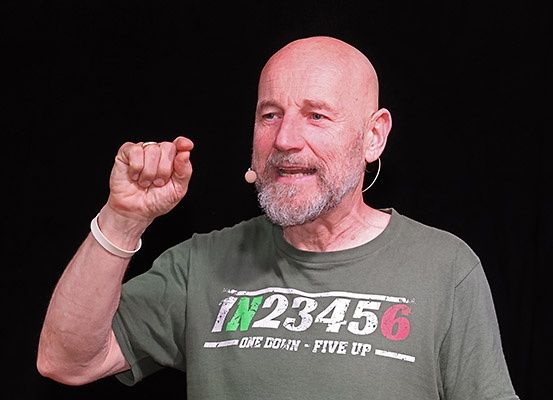 Ein kahlköpfiger Mann mit Bart trägt ein graues T-Shirt mit weißem Text und steht vor schwarzem Hintergrund, er gibt eine Handgeste und lächelt.