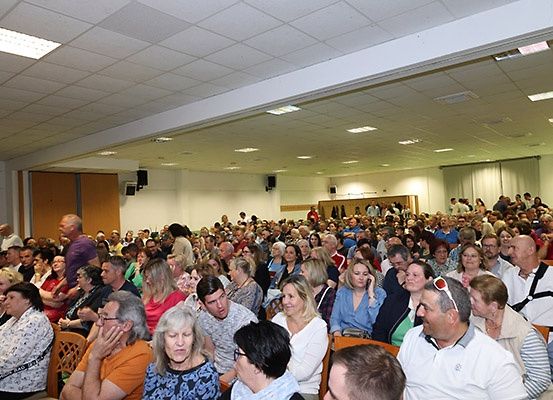 Eine große Anzahl von Menschen sitzt in Stühlen in einem Konferenzsaal, einige tragen Brillen und Hüte. Sie beobachten etwas auf der Bühne, und einige haben ihre Hände auf ihren Gesichtern.