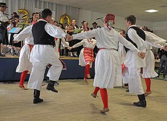 Eine Gruppe von Menschen in traditionellen Kostümen tanzt auf einer Bühne, während Musiker im Hintergrund spielen.