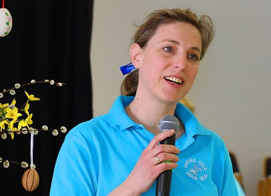 Eine Frau in einem blauen Poloshirt mit Logo spricht in ein Mikrofon. Dahinter befindet sich eine Blumendekoration.