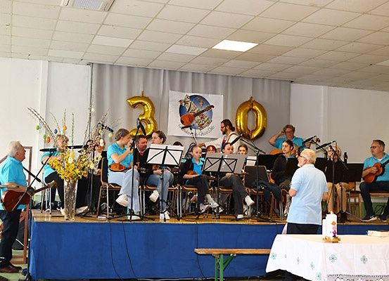 Eine Gruppe von Musikern in blauen Hemden spielt Instrumente auf einer Bühne mit Luftballons und einem Banner zur Feier eines Meilensteins.