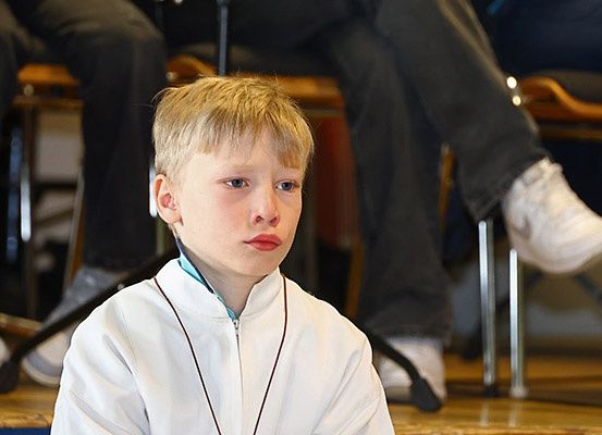 Ein junger Junge mit blonden Haaren sitzt in einem Raum und trägt eine weiße Jacke und eine Halskette. Hinter ihm sitzen zwei Personen auf Stühlen.