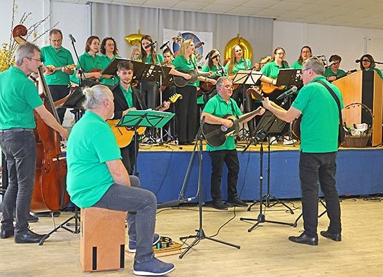 Eine Gruppe von Musikern in grünen Kleidern spielt auf der Bühne mit verschiedenen Instrumenten, darunter Gitarren, Celli und Flöten, während ein Mann vorne singt.