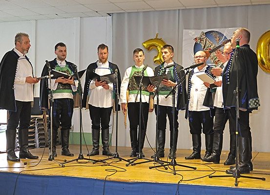 Ein Chor von Männern in traditioneller Kleidung singt auf der Bühne mit Mikrofonen und Notenständern vor ihnen. Hinter ihnen steht ein Banner mit der goldenen Zahl 50.