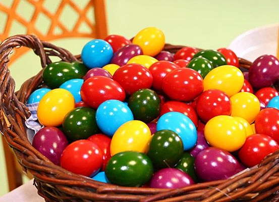 Ein Korb aus Weide gefüllt mit bunten Ostereiern in verschiedenen Farben, auf einem Tisch mit einem gemusterten Stuhl im Hintergrund.