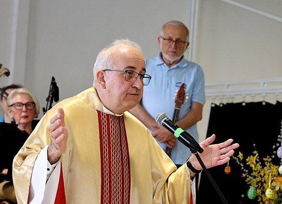 Ein Priester in einem gelben und roten Gewand spricht in ein Mikrofon mit einer Osterdekoration auf der rechten Seite. Hinter ihm steht ein Mann mit einer Gitarre in der Nähe eines schwarzen Vorhangs.