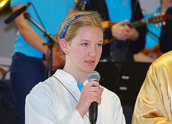 Ein junges Mädchen mit einem blauen Stirnband hält ein Mikrofon und scheint bei einer musikalischen Veranstaltung aufzutreten oder zu sprechen. Dahinter sind verschwommene Figuren von Menschen zu sehen, die Gitarren spielen.