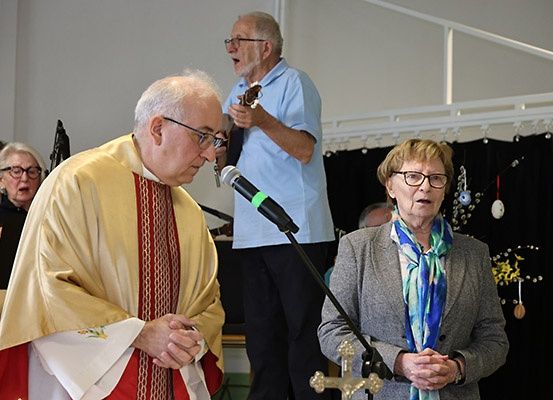 Ein Priester steht vor einem Mikrofon, in gold-roter Kleidung. Neben ihm steht eine Frau mit einem Schal, beide blicken in die Menge. Hinter ihnen spielt ein Mann Gitarre.
