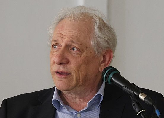 Ein älterer Mann mit weißen Haaren in einem schwarzen Anzug und einem hellblauen Hemd spricht in ein Mikrofon in einem Raum mit einer weißen Wand.