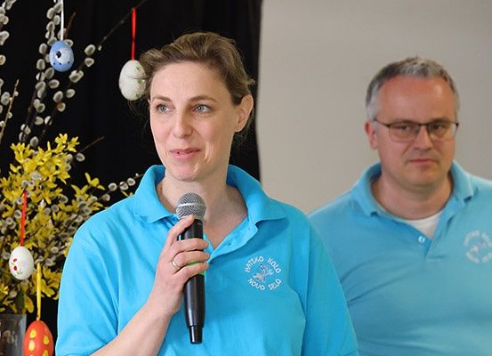 Eine Frau spricht in ein Mikrofon und trägt ein blaues Polohemd mit Logo. Ein älterer Mann steht hinter ihr. Sie befinden sich in einem Raum mit weißer Wand und Osterdekoration.