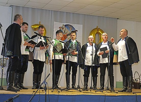 Eine Gruppe von Männern in traditionellen Kostümen singt auf der Bühne mit Mikrofonen. Einige halten Bücher, und einer gestikuliert beim Singen. Hinter ihnen ist ein Banner und Luftballons zu sehen.
