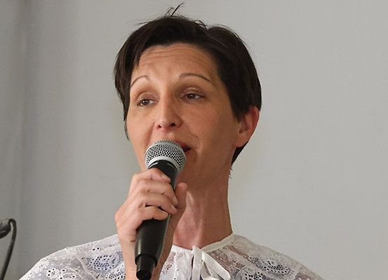 Eine Frau mit kurzen Haaren spricht in ein Mikrofon. Sie trägt ein weißes Oberteil und steht vor einer weißen Wand.