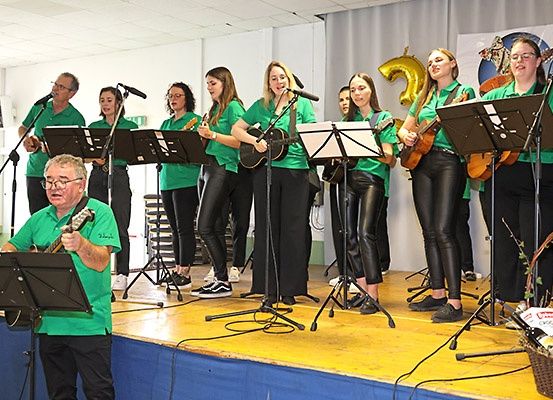 Eine Band aus Frauen und einem Mann auf der Bühne, alle in Grün gekleidet, spielen Gitarren. Der Mann steht vorne, während die Frauen hinter ihm stehen. Sie schauen alle in Richtung Publikum. Hinter ihnen befindet sich ein weißer Hintergrund mit der Nummer 25 und einem Luftballon.