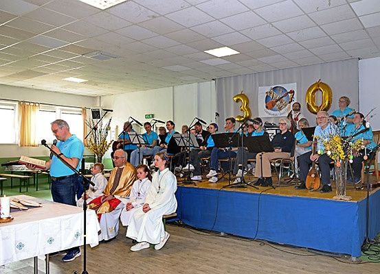 Eine Gruppe von Menschen in blauen Shirts spielt auf der Bühne mit Musikinstrumenten. Ein Mann liest ein Buch auf einem Podium. Ein Priester und zwei Kinder sitzen auf einer Bank. Eine Kerze und Blumen stehen auf der Bühne.