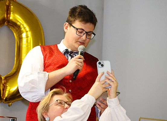 Ein Mann mit Brille und Fliege hält ein Mikrofon, während eine Frau ein Foto mit ihrem Handy macht. Hinter ihnen befindet sich ein goldener Ballon.