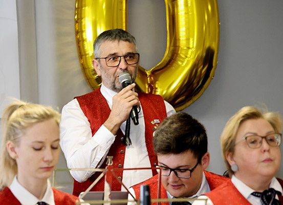 Ein Mann mit Brille spricht in ein Mikrofon, während andere zusehen. Hinter ihm befindet sich ein goldener Ballon in Form der Zahl 50.