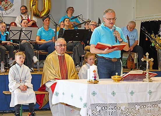 Eine kirchliche Zeremonie findet auf einer Bühne statt. Ein Priester liest aus einem Buch und hält ein Mikrofon. Ein junger Junge in einem weißen Gewand kniet vor einem Tisch mit einer Kerze und einem Kreuz. Dahinter spielt ein Chor Musikinstrumente und singt.