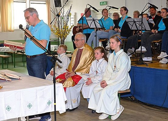 Ein Priester leitet einen Gottesdienst in einer Kirche, wobei Chormitglieder und Teilnehmer sitzen, eine Person ein Buch liest und ein Musiker Gitarre spielt.