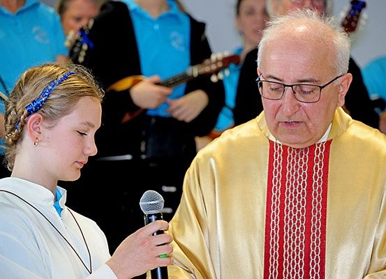 Ein Priester spricht zu einem jungen Mädchen, das ein Mikrofon hält. Hinter ihnen sind andere, einige spielen Gitarre.