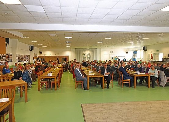 Eine große Gruppe von Menschen sitzt an Tischen in einem Raum mit weißen Decken und grünem Boden. Einige Personen tragen Anzüge, und es hängen mehrere Bilder an den Wänden.